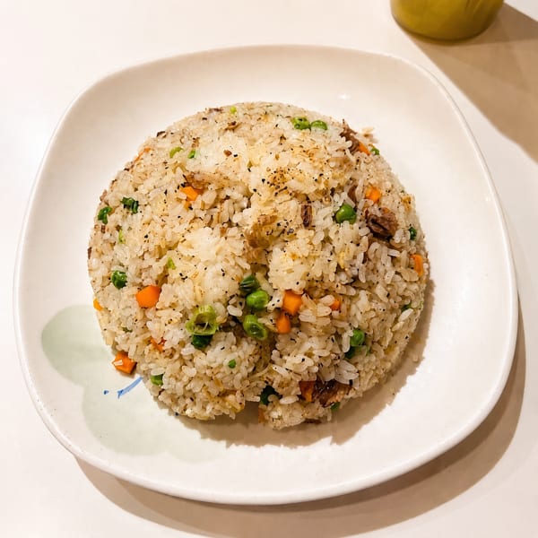 A white plate holds a mound of fried rice with peas, carrots, and meat, sprinkled with pepper.