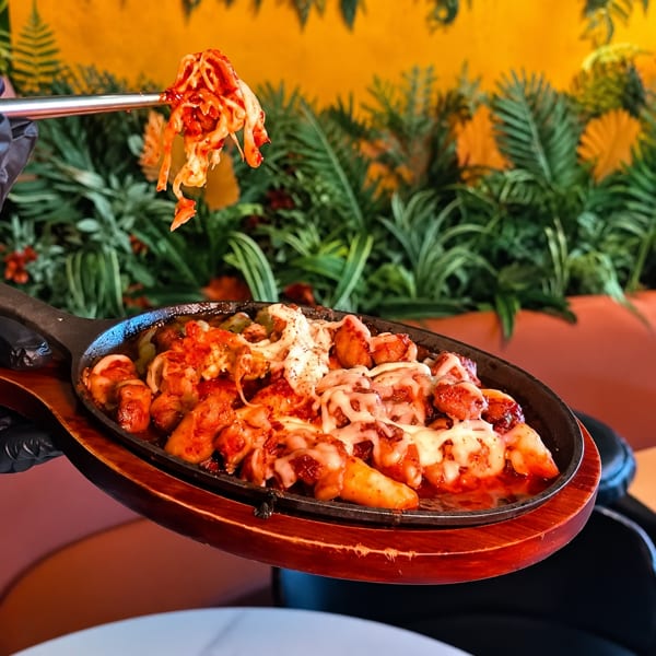 Spicy Korean chicken and rice cakes with melted cheese in a sizzling skillet, with kimchi held by chopsticks.