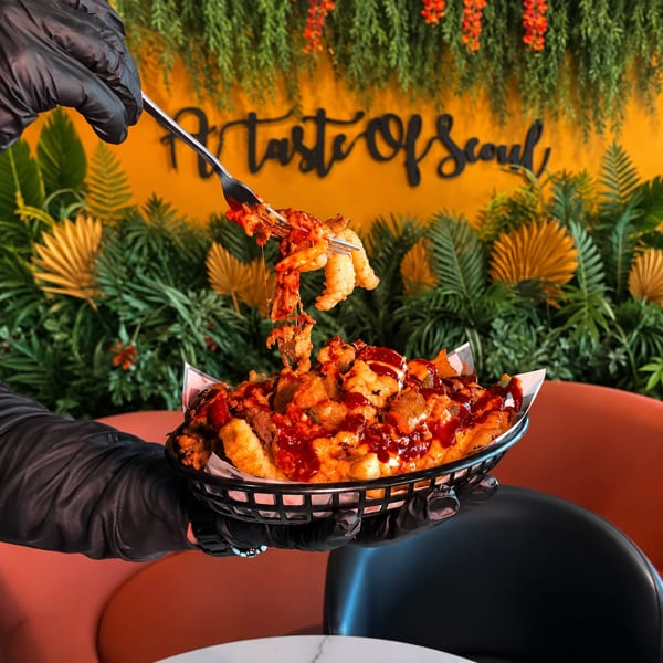 Gloved hand holding a basket of loaded fries with a fork lifting some, against a "Taste Of Seoul" wall.