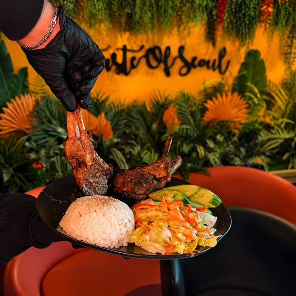 A hand in a black glove holds a plate of lamb chops, rice, and salad at Taste Of Seoul.