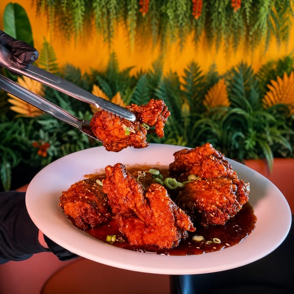Gloved hand using tongs to place a piece of crispy, saucy chicken onto a white plate.